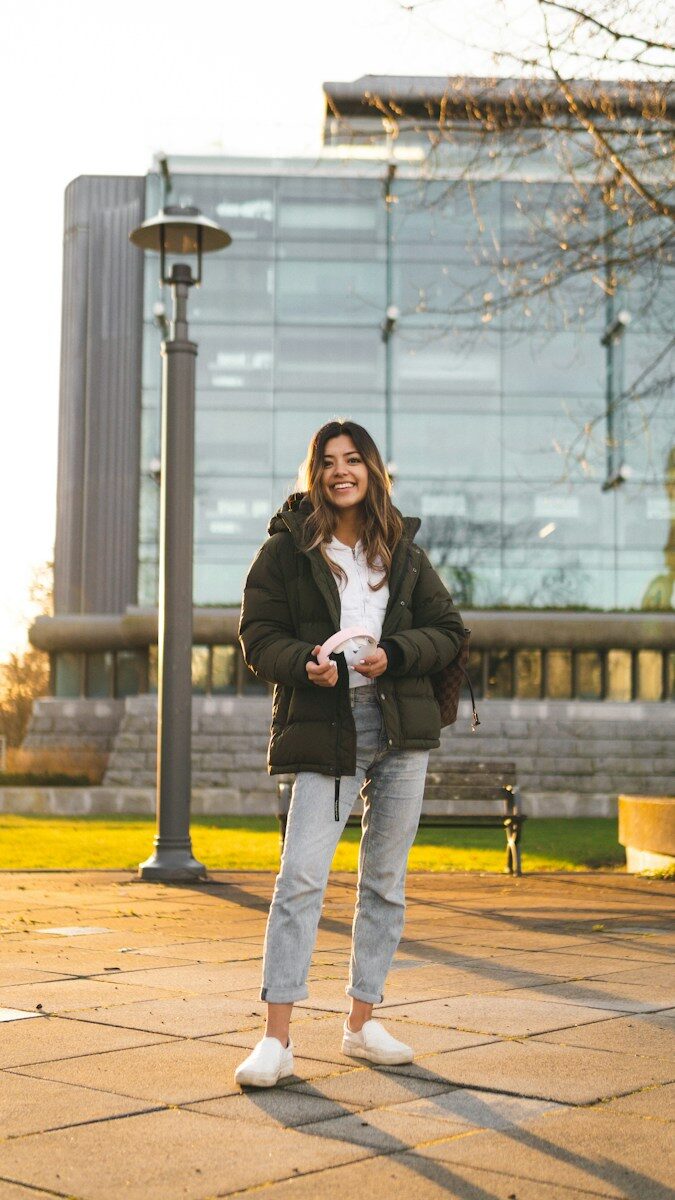 A woman in a dark green puffer jacket holding headphones on a paved plaza